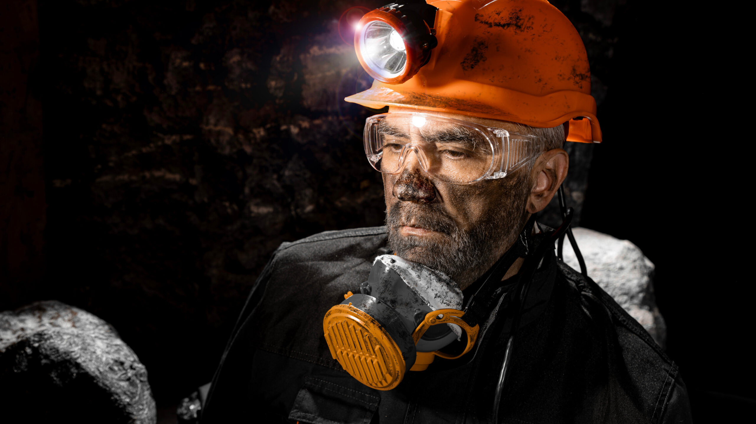 A male underground coal miner covered in black dust wearing a helmet with a flashlight and a respirator around his neck.