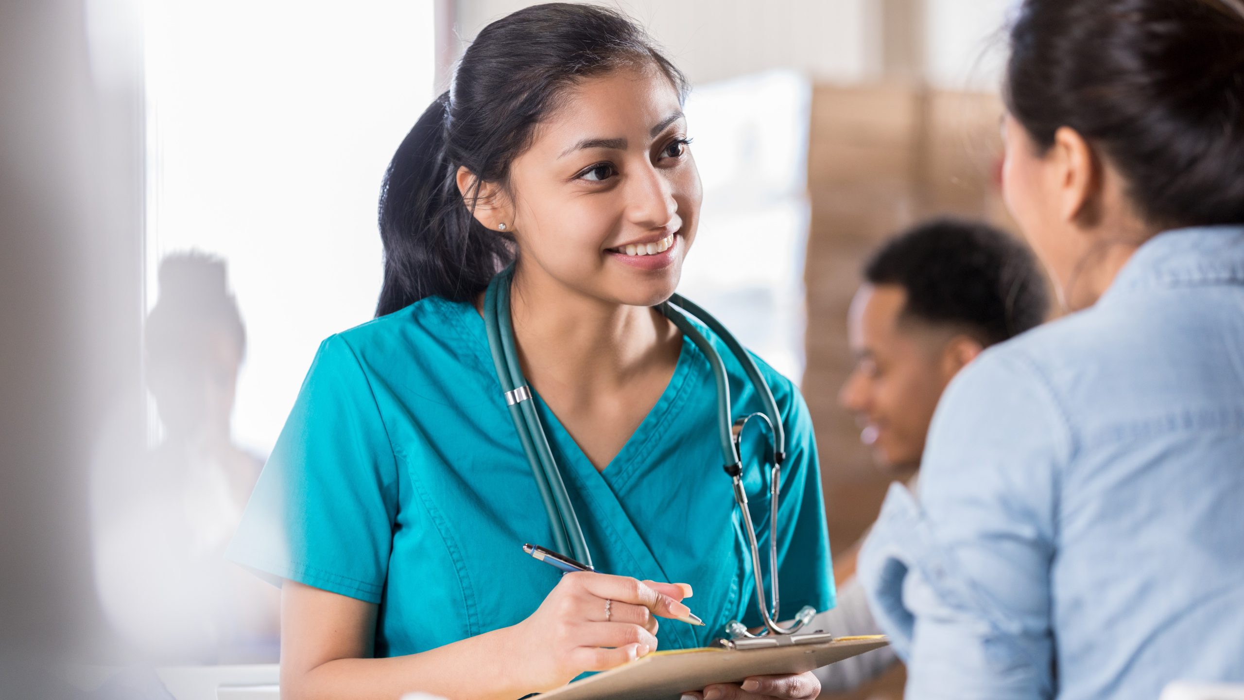 woman wearing blue scrubs and holding a clipboard talking to a person