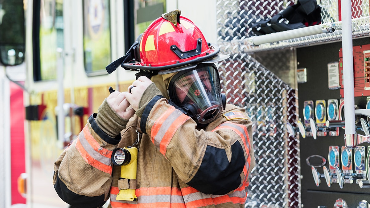 Firefighter in full gear strapping on a respirator in front of a fire truck.