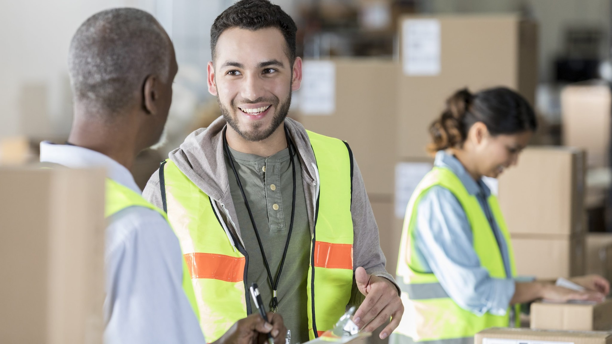 Three warehouse workers wearing yellow safety vests. In the background, a worker affixes a label to a box. In the foreground, a worker is talking to his manager.