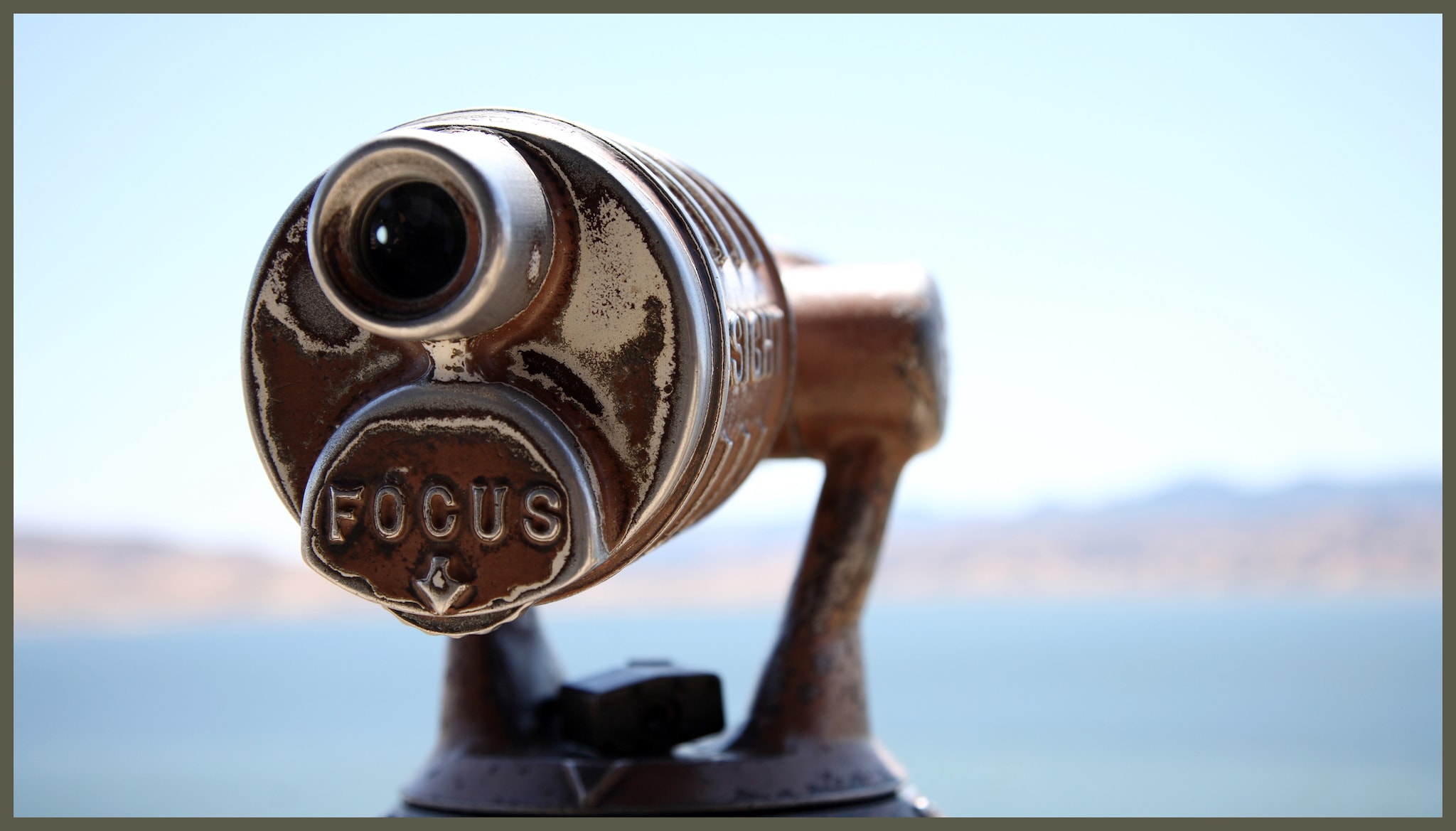 Telescope in foreground with mountain and ocean scenery in background blurred out. View of telescope is where you put your eye to look through and the word focus is shown.