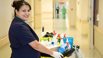 Adult cleaner with cleaning cart in a hospital hallway.