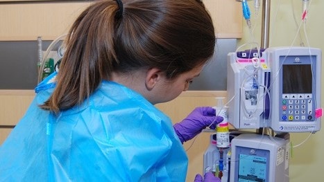 Healthcare worker wearing a gown and gloves administers medication in a syringe through a pump.