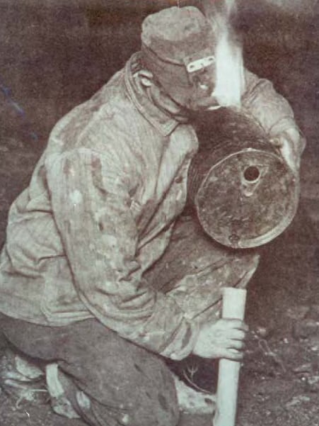 Vintage photograph of miner kneeling and pouring black powder dynamite cartridge while wearing a camp lamp with an open flame.
