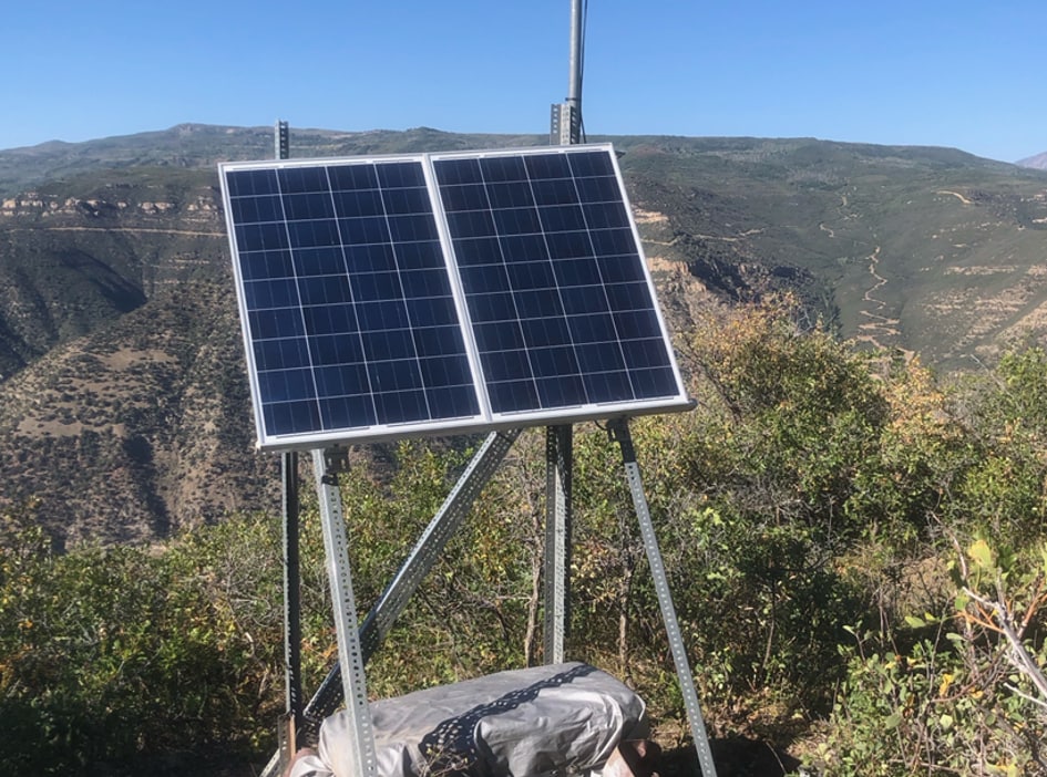 A blue-grey solar panel on metal supports over a grey seismic station vault against a mountain terrain backdrop.