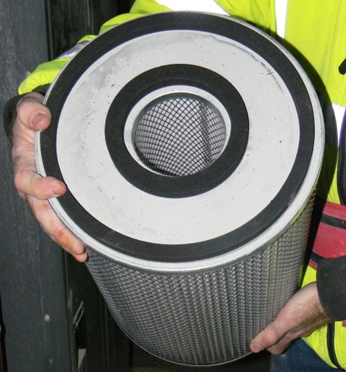 A mine worker holding the filter before it is to be installed in the filter housing, with the angle of the photo showing the double gasket seal near the filter�s smaller inner diameter and filter�s larger outer diameters. The filter is approximately eight inches in diameter and twelve inches long.