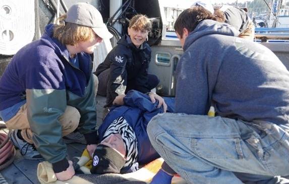 A photo on the deck of a commercial fishing vessel of four people crouched around a prone, unresponsive man simulating an emergency aboard a vessel as part of the Fisherman First Aid Safety Training program.