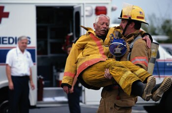 A firefighter carries an older firefighter to an ambulance