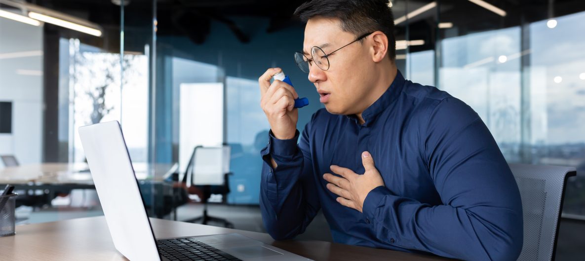 An Asian man office worker having asthma attack at workplace, He sits at a table, uses an inhaler