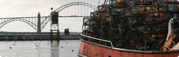 An image with a Dungeness crab vessel loaded with pots in the foreground and the Yaquina Bay Bridge in the background.