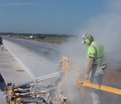 A construction worker in dust while operating a dowel drill on a concrete runway.