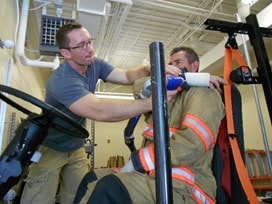 An assistant digitizes a firefighter study participant's wrist location using a Faro Arm device, while the participant reaches for his seatbelt in turnout gear.