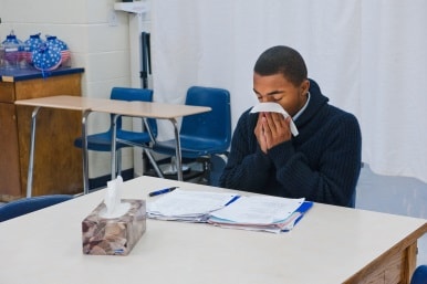 Teen boy at desk in a classroom blowing his nose with a tissue.