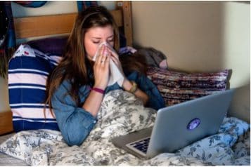 College student blowing her nose with a tissue in bed in her dorm room.