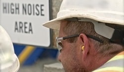 A construction worker wearing hearing protection in front of a sign which reads "High Noise Area"