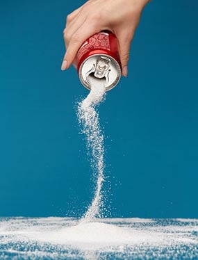 A generic can of soda being held in a pour position, with sugar flowing from it. Children younger than 24 months old should not be given foods or drinks with added sugar.
