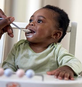 طفل عمره 6 أشهر يجلس على كرسي مرتفع ويتغذى على الأطعمة الصلبة لأول مرة من الملعقة.