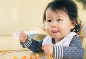 An older infant placing food into her mouth with her hands.