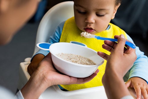 An adult feeding a baby sitting in a high chair a spoonful of oatmeal with a blue spoon