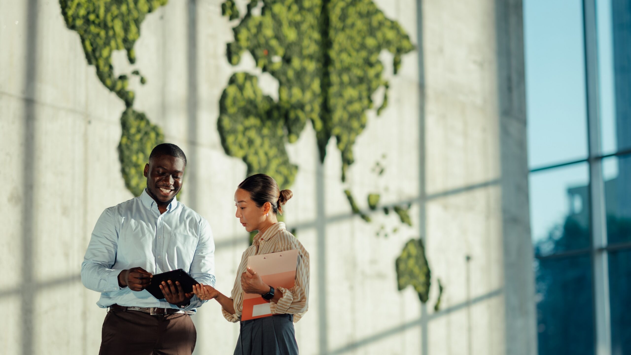 A business man and woman discuss global strategies in a modern office with a large green world map on a background wall in a foyer lit with alot of natural light.