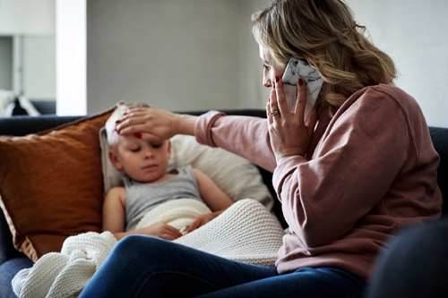 A mother wearing a surgical mask making a phone call while aiding to her sick young son at home