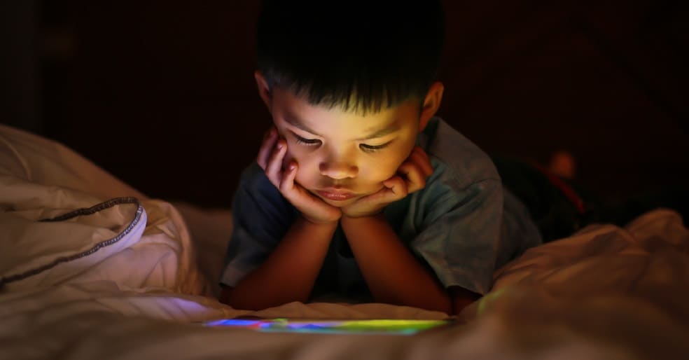 Boy sitting under covers in bed looking at tablet
