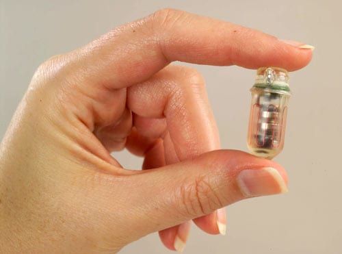 The SmartPill, approximately 1.06 inches long by .46 inches in diameter, held by a person’s thumb and index finger.