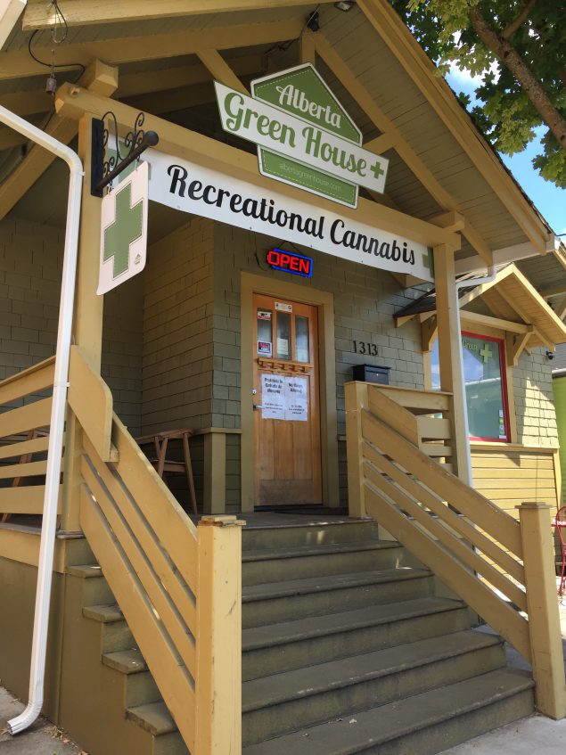 A photograph shows the front of a retail store that sells marijuana. Signs on the front porch read “Alberta Green House” and “Recreational Cannabis”, and there is a sign with a large green plus sign. Signs on the door say “No Minors Allowed” and “Prohibida Entrada de Menores.”