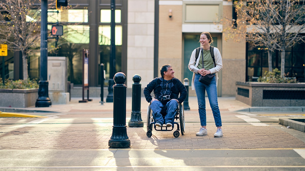 Man in wheelchair and woman standing about to cross the road.