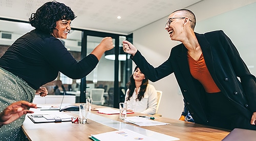 Black businesswoman and white businesswoman fist-bumping across a conference table.