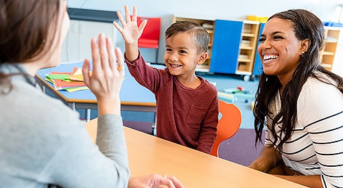 Young, elementary student getting a high-five from his teacher, with mom by his side.