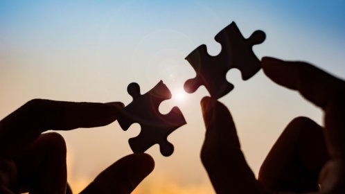 Two hands each holding a puzzle piece in the foreground and a sunset in the background.