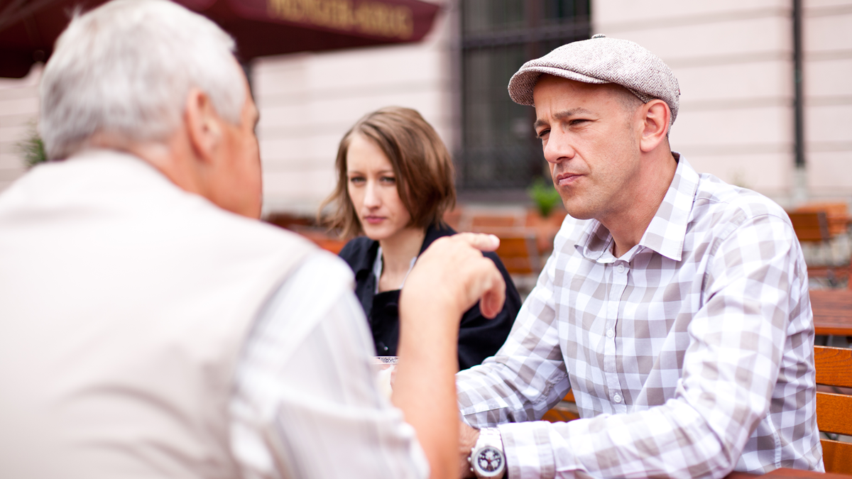 Two men and one woman sitting outdoors at a caf&eacute;; one man is pointing at other man.