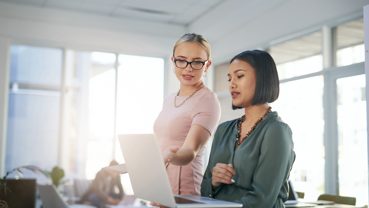 Two businesswomen; one holding a laptop while the other one gestures to something on the screen