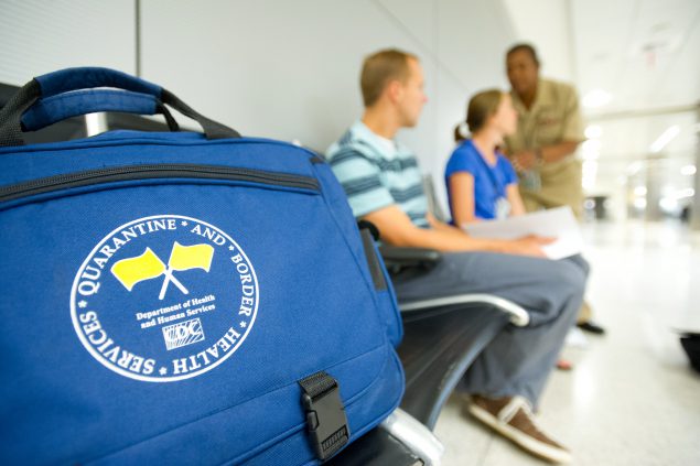 Foreground has a blue bag with the words “Quarantine and Border Health Services” and yellow quarantine flags on it. Blurred in the background is a CDC Quarantine Public Health Officer assessing a sick traveler and companion at a US international airport.