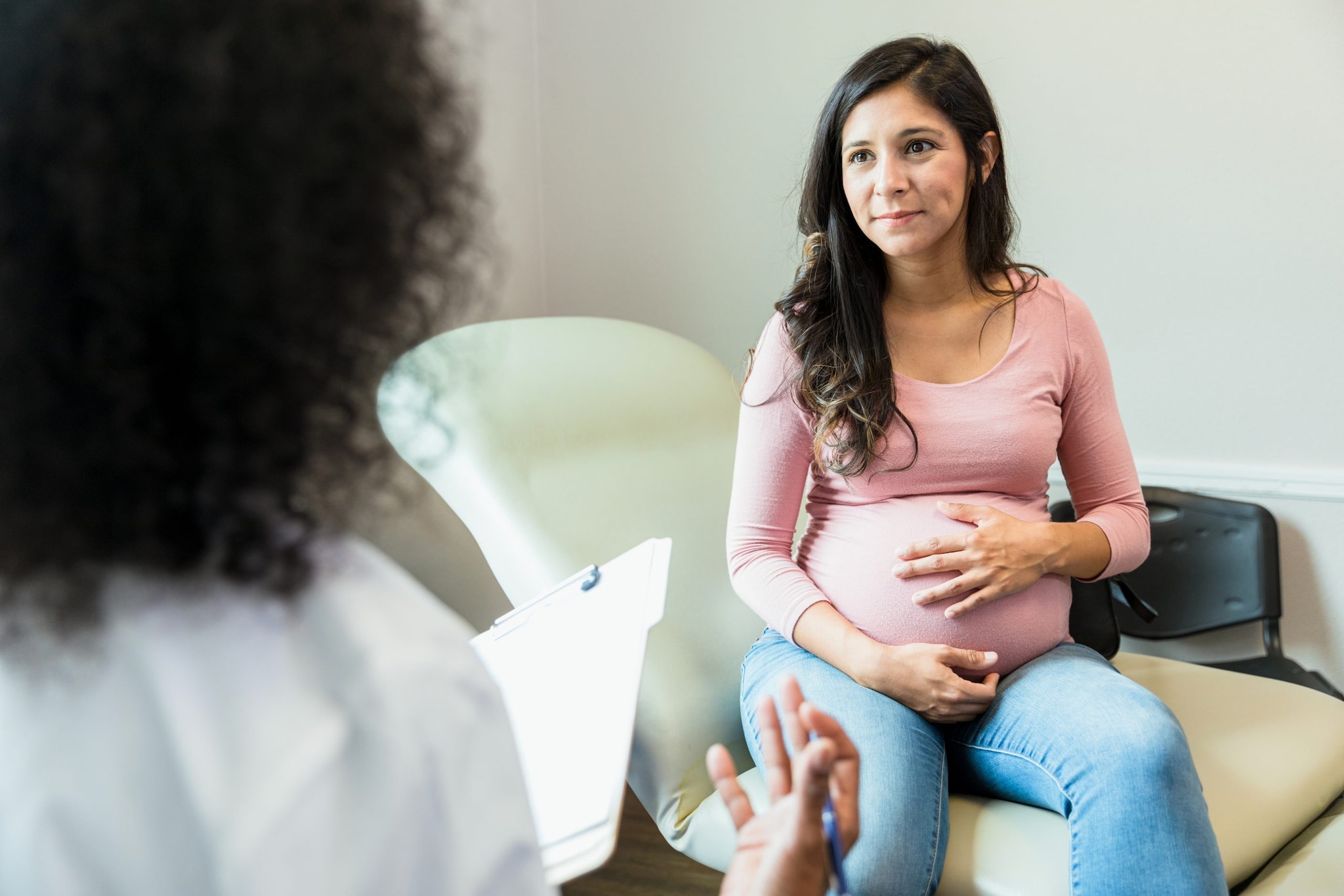 A standing healthcare provider is holding a chart and speaking with a sitting pregnant woman