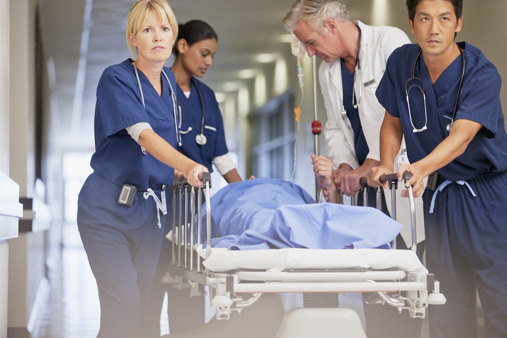 A four-person healthcare team rolling a patient draped in blue on a gurney through a hospital hallway