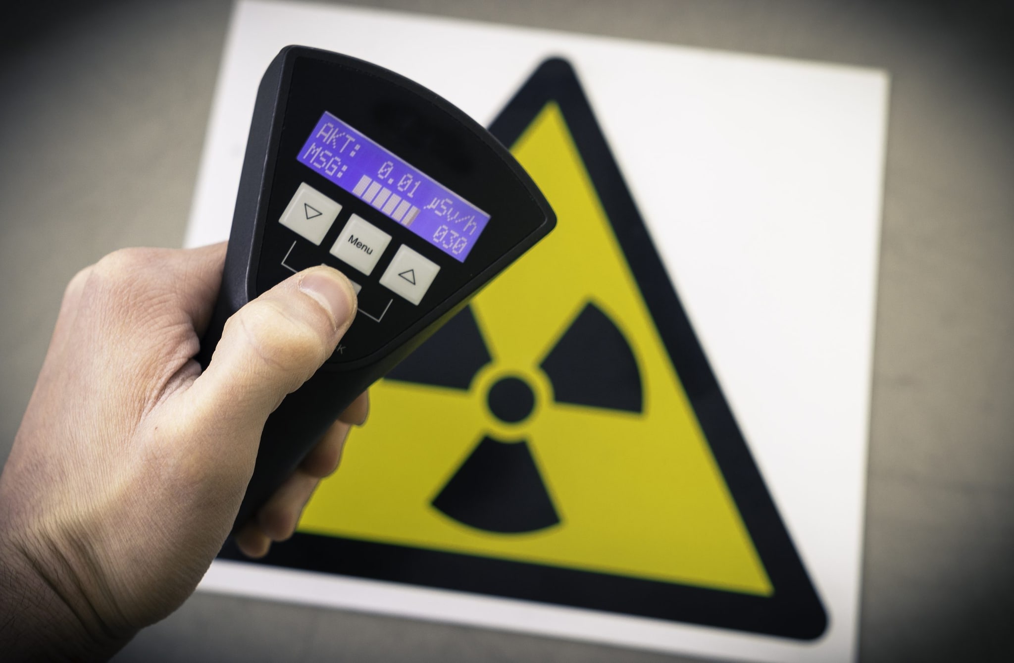 Hand holding device to measure radiation levels. Sign with triangle radiation tri-foil symbol is in the background.