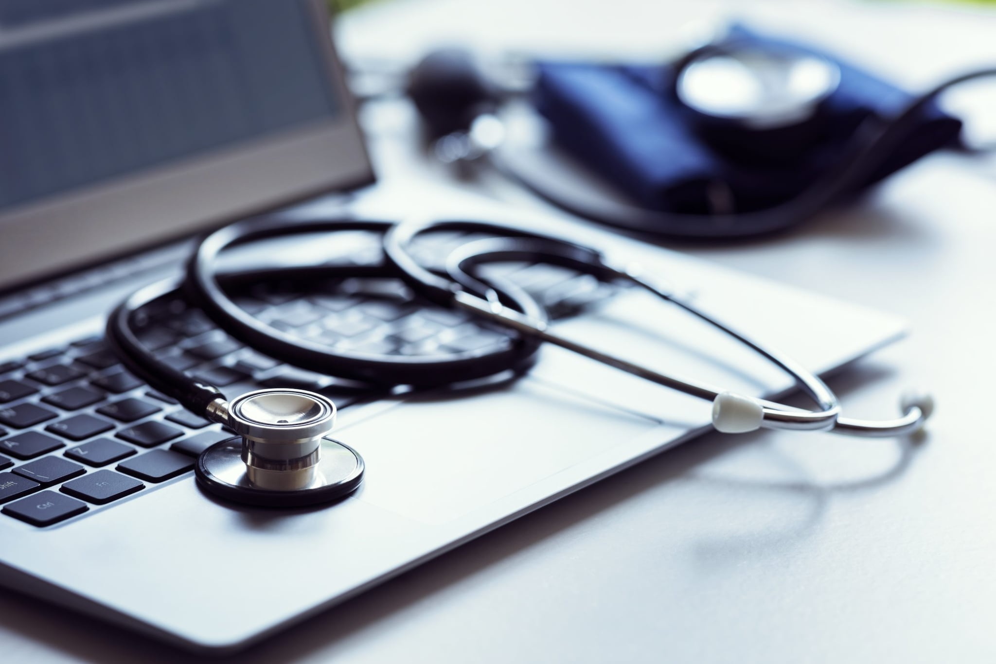 A black stethoscope draped across a laptop keyboard with a blood pressure cuff laying in the background
