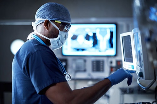 a doctor in a blue surgical gown and mask looking at a monitor with xrays showing in the background