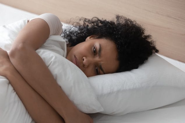 Sad depressed african woman hugging pillow lying in bed alone