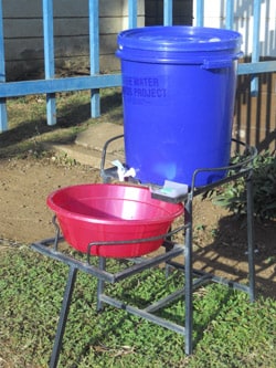 Handwashing station with plastic bucket containing a tap, metal stand, basin for catching water and soap.