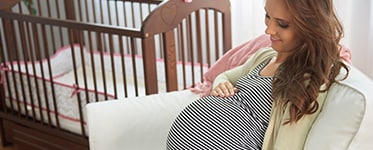 A pregnant woman sitting in front of a crib, smiling at her belly.
