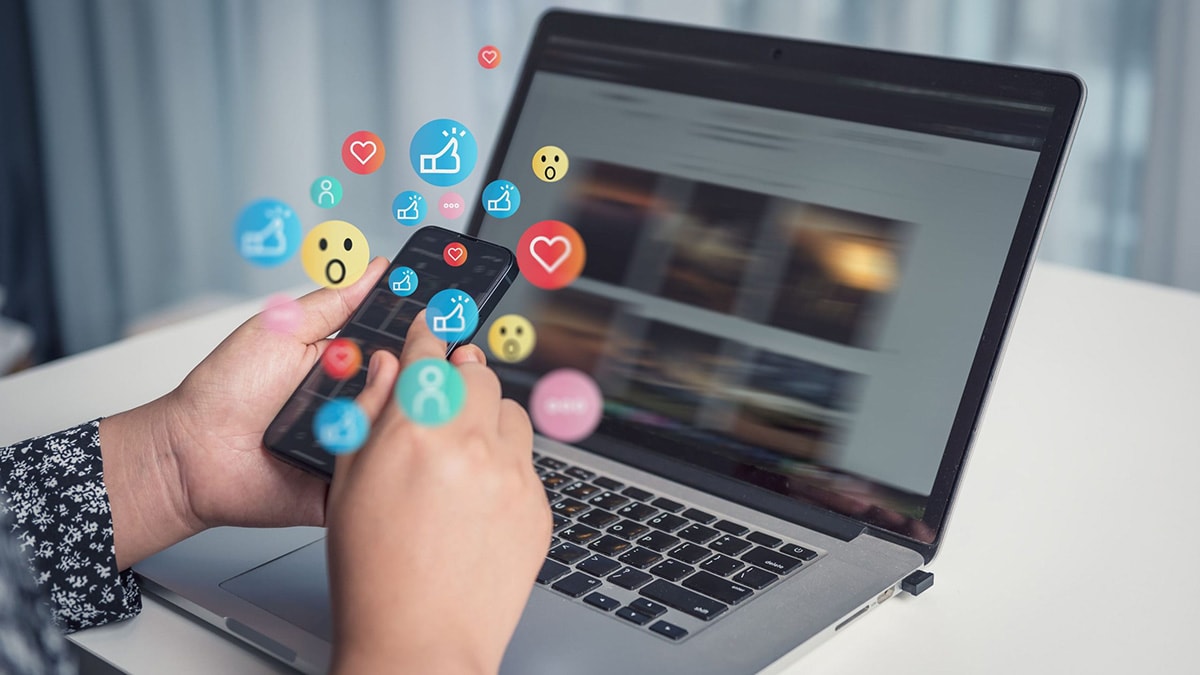Person holding cell phone in front of laptop with a bunch of social media emojis rising up from the phone.