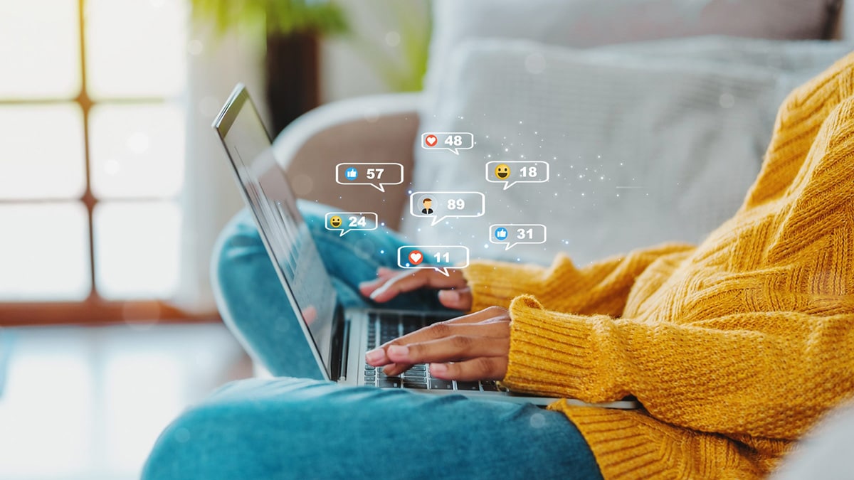 Person wearing a yellow sweater and jeans, sitting on a couch with their laptop open. A bunch of comment bubbles showing social media emojis are above the person's hands over the keyboard.