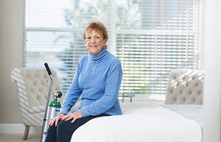 Mujer sentada en una cama usando un tanque de oxígeno portátil.