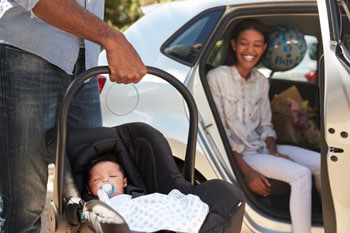 Imagen de un padre que lleva a su bebé en el asiento de seguridad mientras la madre sonríe en el auto. 