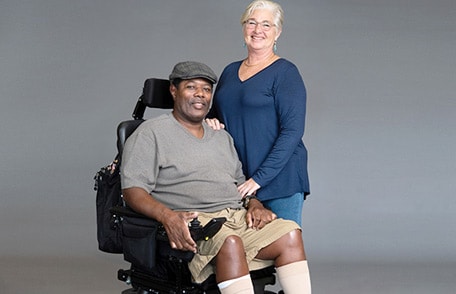 Paciente con esclerosis lateral amiotrófica (ELA) en silla de ruedas sonriendo con un cuidador.