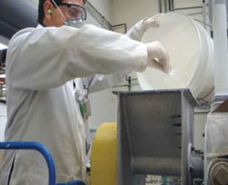 Un trabajador que labora en la producción de nanopartículas viste un equipo de protección dérmico, respiratorio y auditivo y un aparato de muestreo personal de aire durante una operación de vertimiento.