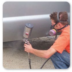 Vista lateral de un trabajador en cuclillas usando protección respiratoria mientras aplica pintura en el costado de un camión